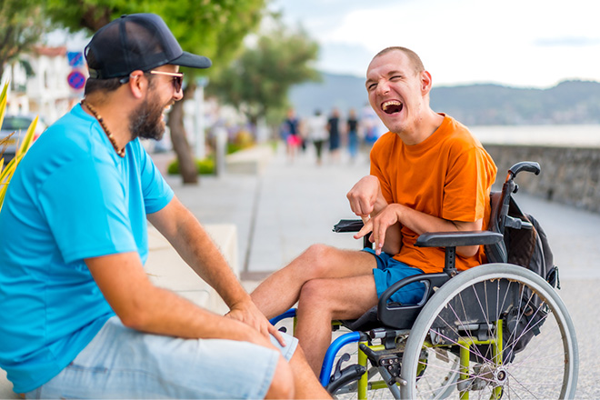 Two men outside on a sunny day, sitting facing each other and laughing happily. One man is wearing a cap and sunglasses, the other man is in a wheelchair. 
